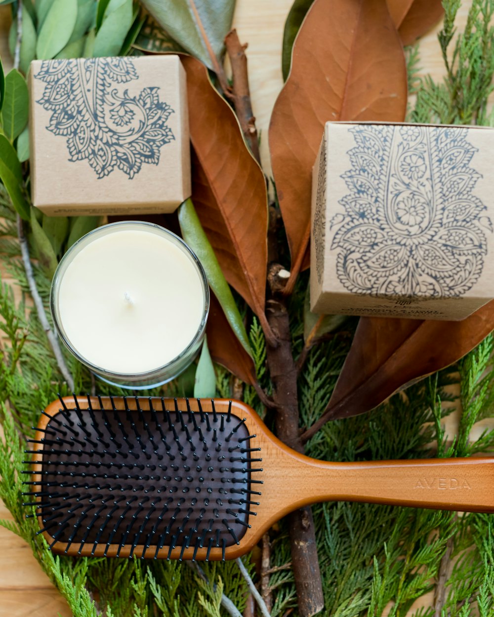 A wooden paddle brush and a candle resting on cedar sprigs and dried magnolia leaves