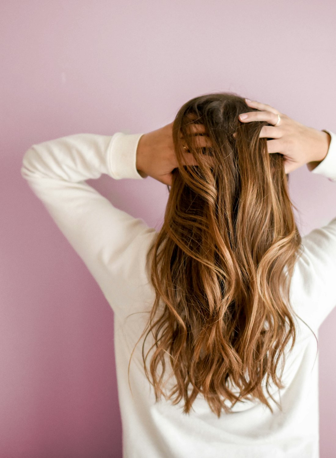 Long honey-brown balayage with soft waves, seen from behind against a blush wall
